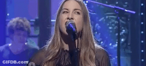 A Woman Is Singing Into A Microphone On A Stage With A Man Playing A Piano In The Background . GIF