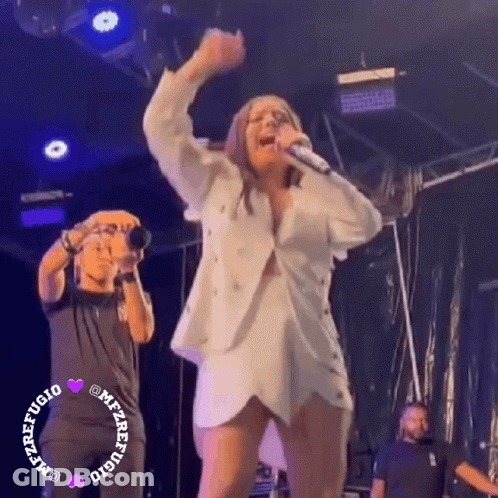 A Woman Is Singing Into A Microphone On Stage While A Man Takes A Picture Of Her . GIF