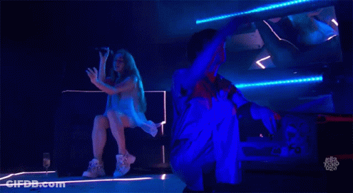 A Woman Is Singing Into A Microphone While A Man Is Kneeling Down Behind Her GIF