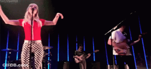 A Woman Is Singing Into A Microphone While A Man Plays A Guitar On A Stage . GIF