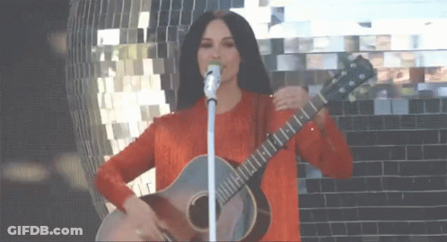 A Woman Is Singing Into A Microphone While Holding A Guitar GIF