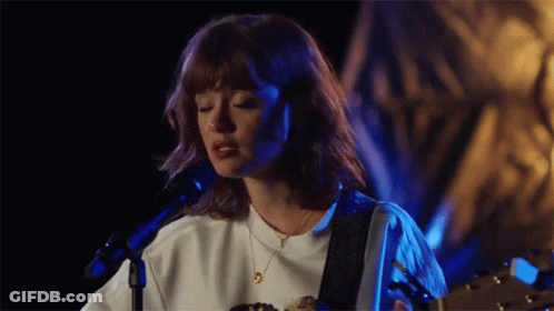 A Woman Is Singing Into A Microphone While Holding A Guitar . GIF