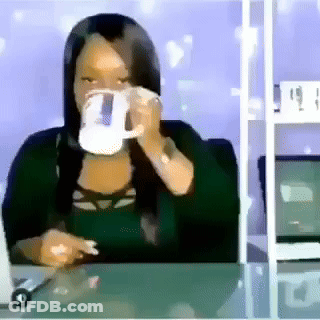 A Woman Is Sitting At A Desk Drinking From A Mug GIF