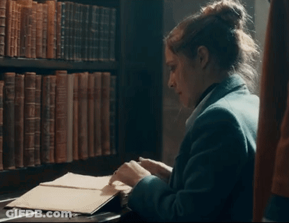 A Woman Is Sitting At A Desk In Front Of A Bookshelf In A Library Looking At A Book . GIF