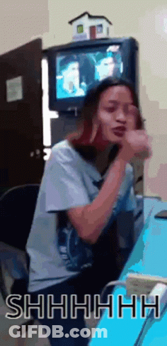 A Woman Is Sitting At A Desk In Front Of A Television And Making A Shhh Gesture GIF