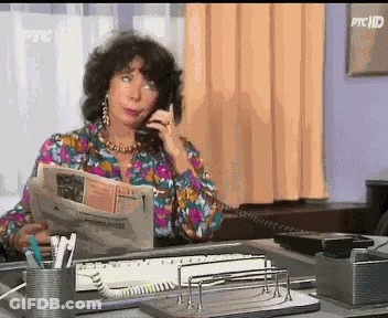 A Woman Is Sitting At A Desk Talking On A Phone And Reading A Newspaper GIF