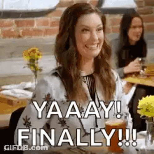 A Woman Is Sitting At A Table Drinking A Beer And Smiling While Another Woman Looks On . GIF