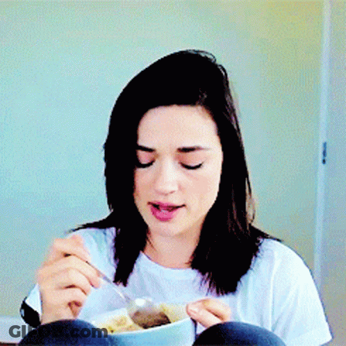 A Woman Is Sitting At A Table Eating A Bowl Of Food With A Spoon GIF