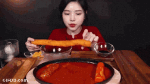 A Woman Is Sitting At A Table Eating A Large Piece Of Food GIF