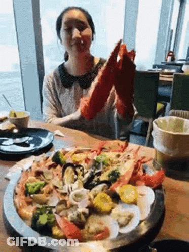 A Woman Is Sitting At A Table Eating A Pizza With Seafood And Vegetables . GIF