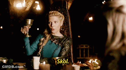 A Woman Is Sitting At A Table Holding A Goblet And The Word Skol Is Next To Her GIF
