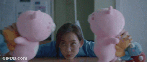 A Woman Is Sitting At A Table Holding Two Pink Pig Stuffed Animals GIF