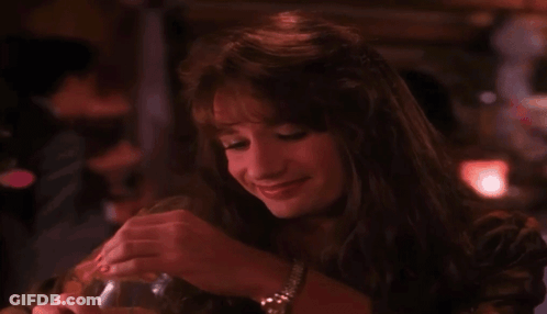 A Woman Is Sitting At A Table In A Dark Room Holding A Glass And Smiling . GIF