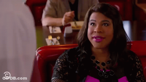A Woman Is Sitting At A Table In A Diner Wearing A Pink Top And Black Lace Top . GIF