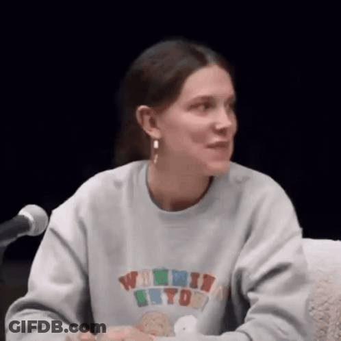 A Woman Is Sitting At A Table In Front Of A Microphone Wearing A Sweatshirt And Earrings . GIF