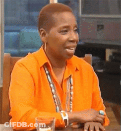 A Woman Is Sitting At A Table Wearing An Orange Shirt And Smiling . GIF