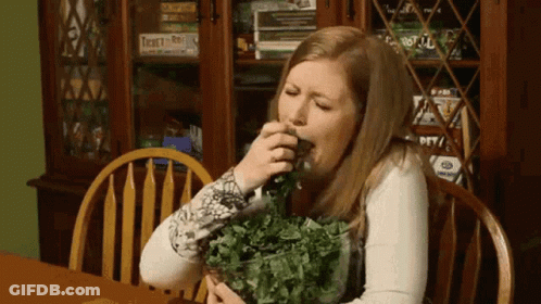 A Woman Is Sitting At A Table With A Bowl Of Greens In Front Of A Bookcase With Twenty-One On It GIF