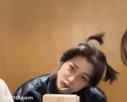 A Woman Is Sitting At A Table With A Box Of Food In Her Hand And Her Hair In A Ponytail . GIF