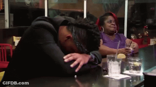 A Woman Is Sitting At A Table With A Drink In Her Hand And A Man Is Laying On The Table With His Head Down . GIF