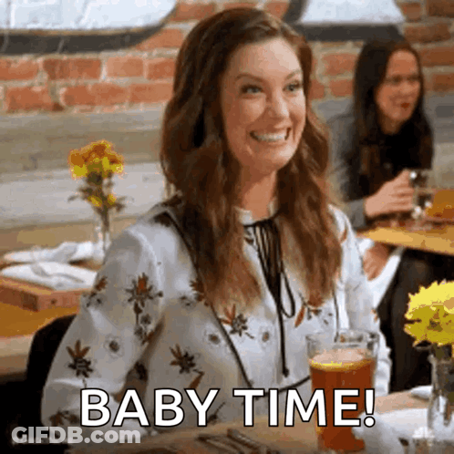 A Woman Is Sitting At A Table With A Glass Of Beer And Smiling With The Words Baby Time . GIF