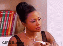 A Woman Is Sitting At A Table With A Glass Of Wine And A Piece Of Food In Her Hand . GIF