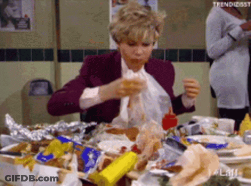 A Woman Is Sitting At A Table With A Lot Of Food On It Eating A Sandwich . GIF