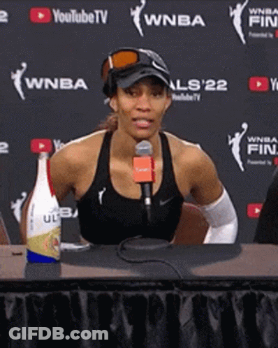 A Woman Is Sitting At A Table With A Microphone In Front Of A Wall That Says Wnba GIF