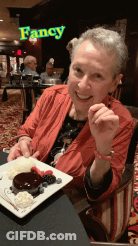 A Woman Is Sitting At A Table With A Plate Of Dessert In Front Of A Sign That Says Fancy GIF