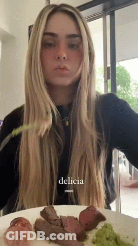 A Woman Is Sitting At A Table With A Plate Of Food And The Word Delicia Is On The Bottom Of The Picture GIF