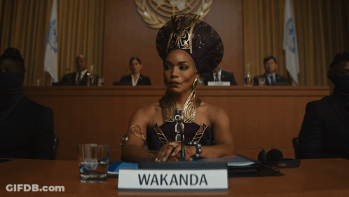 A Woman Is Sitting At A Table With A Sign That Says Wakanda GIF