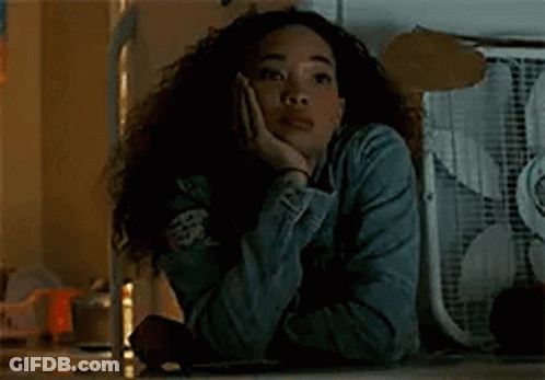 A Woman Is Sitting At A Table With Her Hand On Her Face And A Fan In The Background . GIF