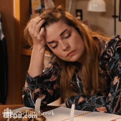 A Woman Is Sitting At A Table With Her Head Resting On Her Hand And The Word Schitts Creek Is On The Bottom Right GIF