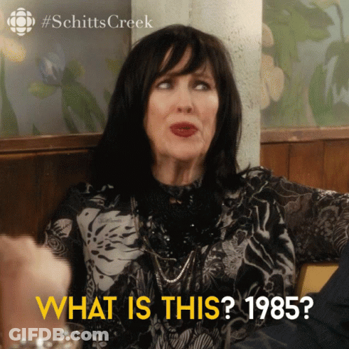 A Woman Is Sitting At A Table With The Words What Is This 1985 On The Bottom GIF