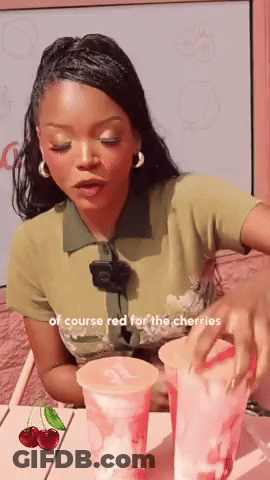 A Woman Is Sitting At A Table With Two Cups Of Cherry Juice GIF