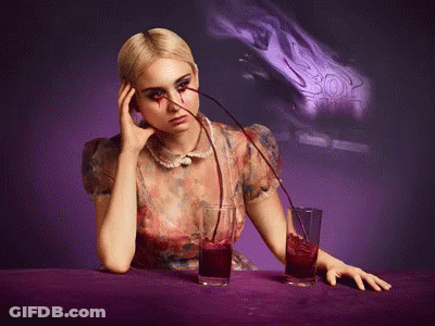 A Woman Is Sitting At A Table With Two Glasses Of Red Liquid Being Poured Into Her GIF
