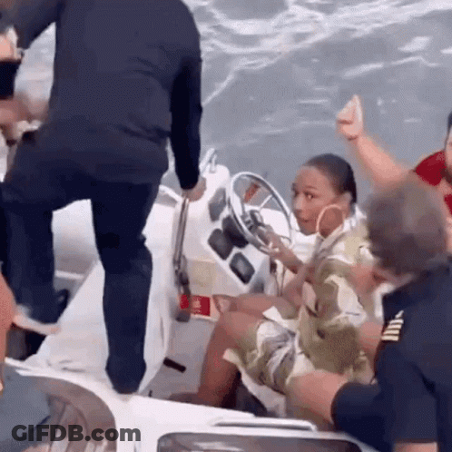 A Woman Is Sitting At The Steering Wheel Of A Boat While A Man Stands Next To Her . GIF
