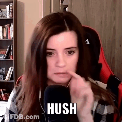 A Woman Is Sitting In A Chair In Front Of A Microphone And Making A Hush Gesture . GIF
