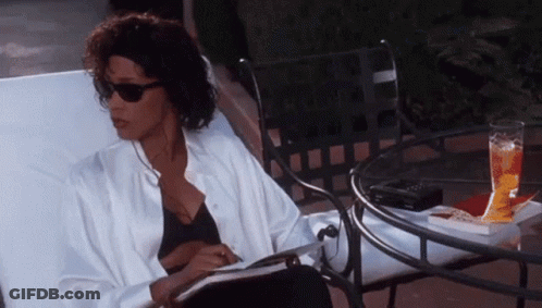 A Woman Is Sitting In A Chair Reading A Book Next To A Table . GIF