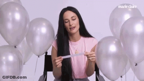 A Woman Is Sitting In A Chair Surrounded By Silver Balloons And Holding A Piece Of Paper . GIF