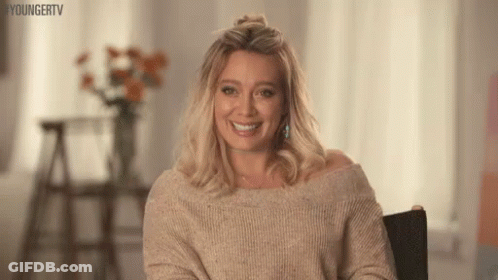 A Woman Is Sitting In A Chair Wearing A Sweater And Smiling . GIF