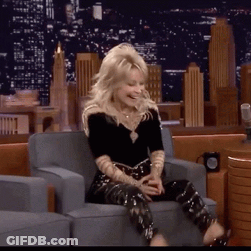 A Woman Is Sitting In A Chair With Her Legs Crossed And Laughing GIF