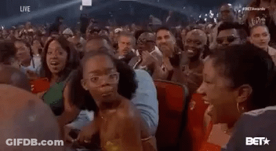 A Woman Is Sitting In A Crowd Of People At A Concert And Making A Funny Face . GIF