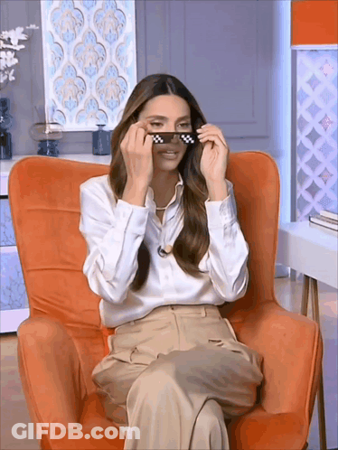 A Woman Is Sitting In An Orange Chair And Wearing Sunglasses GIF