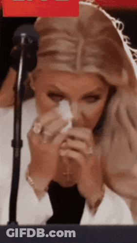 A Woman Is Sitting In Front Of A Microphone Holding A Napkin To Her Nose GIF