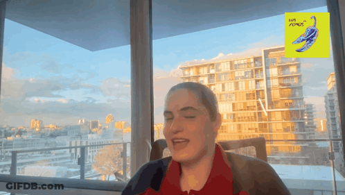A Woman Is Sitting In Front Of A Window With A Yellow Sign That Says Love Plumes On It GIF