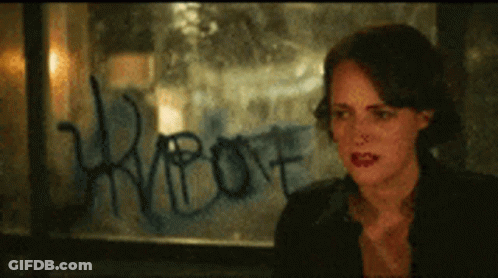 A Woman Is Sitting In Front Of A Window With Graffiti On It That Says ' Riot ' GIF