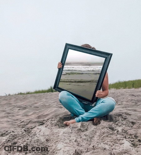 A Woman Is Sitting In The Sand Holding A Picture Frame With A Beach Scene In It GIF