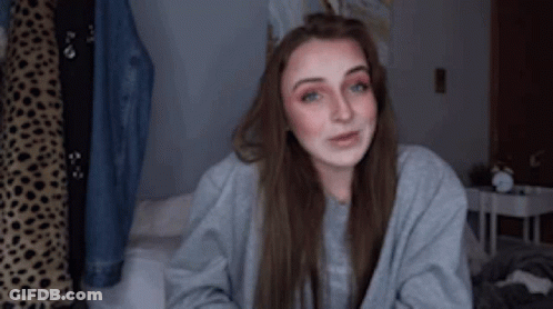 A Woman Is Sitting On A Bed Wearing A Grey Sweater And Smiling . GIF