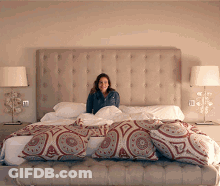 A Woman Is Sitting On A Bed With A Headboard And Pillows GIF