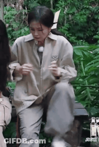 A Woman Is Sitting On A Bench In The Woods Holding A Bottle . GIF